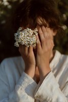 Young woman covering face with hands holding small white daisy flowers in natural outdoor setting with soft warm lighting for wellness and mindfulness concepts.