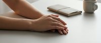 Closeup of female hands resting on a white desk with a notebook and cup in the background, conveying calm and focus.