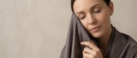 Young woman with closed eyes holding soft gray fabric against her face, enjoying tactile comfort and relaxation in minimalist neutral setting.