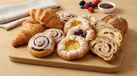 Assorted fresh pastries and baked goods on wooden board including croissants, cinnamon rolls, fruit danishes and artisan bread for breakfast bakery display.