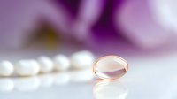 Rose gold wedding ring on white surface with purple orchid flowers in soft focus background creating romantic bridal jewelry display for engagement concepts.