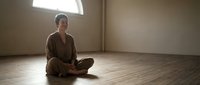 Young Asian woman meditating in empty room with natural light streaming through window. Peaceful mindfulness practice in minimalist space.