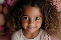 Mixed race child with bright blue eyes and natural curly hair smiling warmly at camera against pink floral background, wearing light colored top.