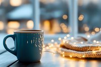 Cozy teal mug with warm knitted blanket and fairy lights creating hygge atmosphere on wooden table. Winter comfort and relaxation concept for home decor.