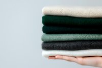 Stack of folded knitted sweaters in neutral and green tones held by female hand against gray background. Cozy winter clothing collection for seasonal wardrobe.