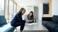 Two professional women reviewing documents at modern office table with large windows and contemporary furniture for business meeting collaboration.