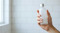 Hand holding white cosmetic bottle with silver cap against blurred bathroom background, suitable for skincare, beauty product marketing, and wellness advertising campaigns.