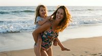 Happy mother and daughter enjoying piggyback ride on sunny beach during golden hour vacation time together creating joyful family memories by ocean waves.