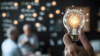 Glowing light bulb with digital network connections held by hand against blurred business meeting background, representing innovation and ideas.