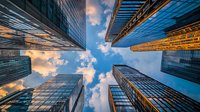 Modern glass skyscrapers reaching into blue sky with fluffy clouds at sunset, upward perspective view showing reflective windows and architectural details.