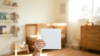 Mature adult male holding blank white card in warm wooden bedroom interior with natural sunlight, suitable for advertising and promotional campaigns.