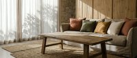 Modern living room interior with wooden coffee table, neutral sofa with colorful pillows, natural lighting through large windows creating warm atmosphere.
