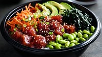Fresh poke bowl with glazed tuna, edamame beans, carrots, avocado and seaweed on black plate, topped with white sesame seeds and green onions.