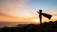 Silhouetted child wearing superhero cape standing on rocky cliff at sunset overlooking ocean, arms outstretched in powerful pose, inspiring courage and imagination for motivational content.