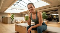 Young woman, Caucasian ethnicity, fitness instructor or wellness professional, holding green smoothie in modern yoga studio with natural light and plants, promoting healthy lifestyle and nutrition.