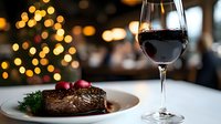 Grilled beef steak with red wine glass on elegant table setting with warm bokeh lights creating festive dining atmosphere for holiday celebration.