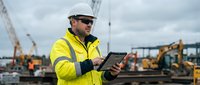 Construction worker in safety gear using digital tablet at industrial site with heavy machinery and cranes in background for project management.