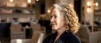 Mature blonde Caucasian businesswoman with curly hair smiling confidently in modern office or restaurant setting with warm ambient lighting.