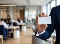 Businessman in dark suit holding blank white business card in modern office conference room with blurred colleagues in background, suitable for corporate branding and professional networking materials.