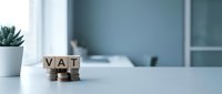 Wooden blocks spelling VAT stacked on white desk with succulent plant and blurred office background, representing tax compliance and business accounting concepts.