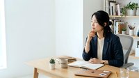 Woman in business casual attire sitting at wooden desk with laptop, notebook, and plant, contemplating work tasks in bright modern office environment.