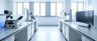 Modern laboratory interior with microscopes and scientific equipment on white countertops. Clean research facility with large windows and monitors for analysis work.