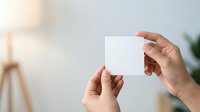 Close-up of two hands holding blank white business card in modern office environment with natural lighting and blurred background for branding mockup.