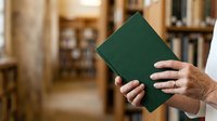 Senior adult with light skin tone holding closed green leather-bound book in library setting with blurred bookshelves background for education and reading concepts.