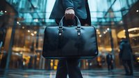 Professional holding elegant black leather briefcase against modern glass building entrance with bokeh lights, urban business lifestyle concept.