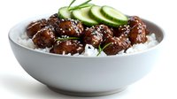 Asian meatballs with sesame seeds served on white rice in ceramic bowl, garnished with fresh cucumber slices and green onions on white background.