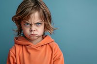 Young child with intense blue eyes wearing orange hoodie showing angry, grumpy expression against blue background, depicting childhood emotions and behavioral concepts.