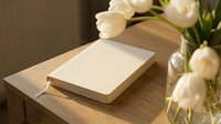 Closed cream notebook on wooden table beside white tulips in glass vase, warm natural light, cozy home office setting.