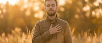 Man with hand on chest standing in golden wheat field during sunset, expressing gratitude or emotion outdoors.