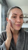 Woman applying exfoliating face scrub in modern bathroom wearing gray bathrobe for skincare routine and spa treatment at home.