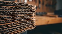Stack of corrugated cardboard sheets showing detailed fluted structure in warehouse setting for packaging and shipping industry applications.