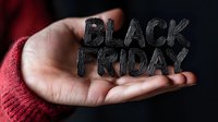 Hand holding charcoal-textured Black Friday text against dark background, wearing red knitted sleeve. Dramatic lighting emphasizes seasonal shopping concept.