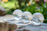 Crystal clear glass spheres on stone surface with natural green bokeh background, macro photography showing reflections and light play through transparent orbs.