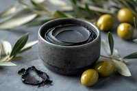 Black charcoal face mask in concrete bowl with fresh green olives and olive branches on gray stone background, natural skincare and spa concept.