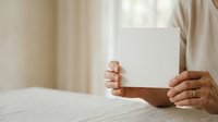Person of medium skin tone holding blank white card or paper in both hands at table with neutral beige background, suitable for mockups, advertising, and promotional campaigns.