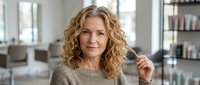 Middle-aged Caucasian woman with curly blonde hair in modern kitchen interior holding cooking utensil with confident expression for culinary lifestyle content.
