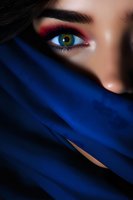 Close-up of green eye with red eyeshadow makeup and dark eyebrow, face partially covered with deep blue fabric creating mysterious middle eastern style look.