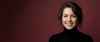 Young professional woman with brown wavy hair smiling confidently in black turtleneck against burgundy background for business and corporate use.