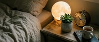 Cozy bedroom nightstand with glowing moon lamp, succulent plant, alarm clock and coffee mug creating warm ambient lighting for relaxation and sleep.