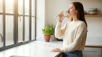 Young woman with long dark hair and light skin tone drinking water from a glass while standing at kitchen counter with potted plant and window in background.