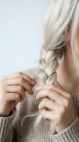 Woman with long platinum blonde hair braiding her hair, wearing a beige knit sweater, shown from behind with hands visible styling the braid for hair care and beauty content.