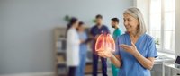 Middle-aged Caucasian female doctor in blue scrubs examining glowing anatomical lung model in modern medical clinic with healthcare team background.