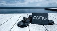 Flip flops and a slate sign reading Vacation on a wooden dock by the ocean, evoking summer travel and relaxation.