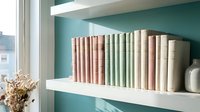Organized bookshelf with neutral colored books on white floating shelves against teal wall creating modern minimalist home library interior design.
