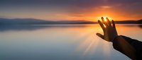 Hand reaching toward golden sunset over calm water creating peaceful spiritual moment with warm light rays and serene lake reflection.