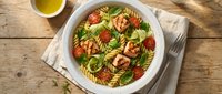 Fresh pasta salad with spiral noodles, cherry tomatoes, basil leaves and herbs in white bowl on rustic wooden table with olive oil for healthy meal.
